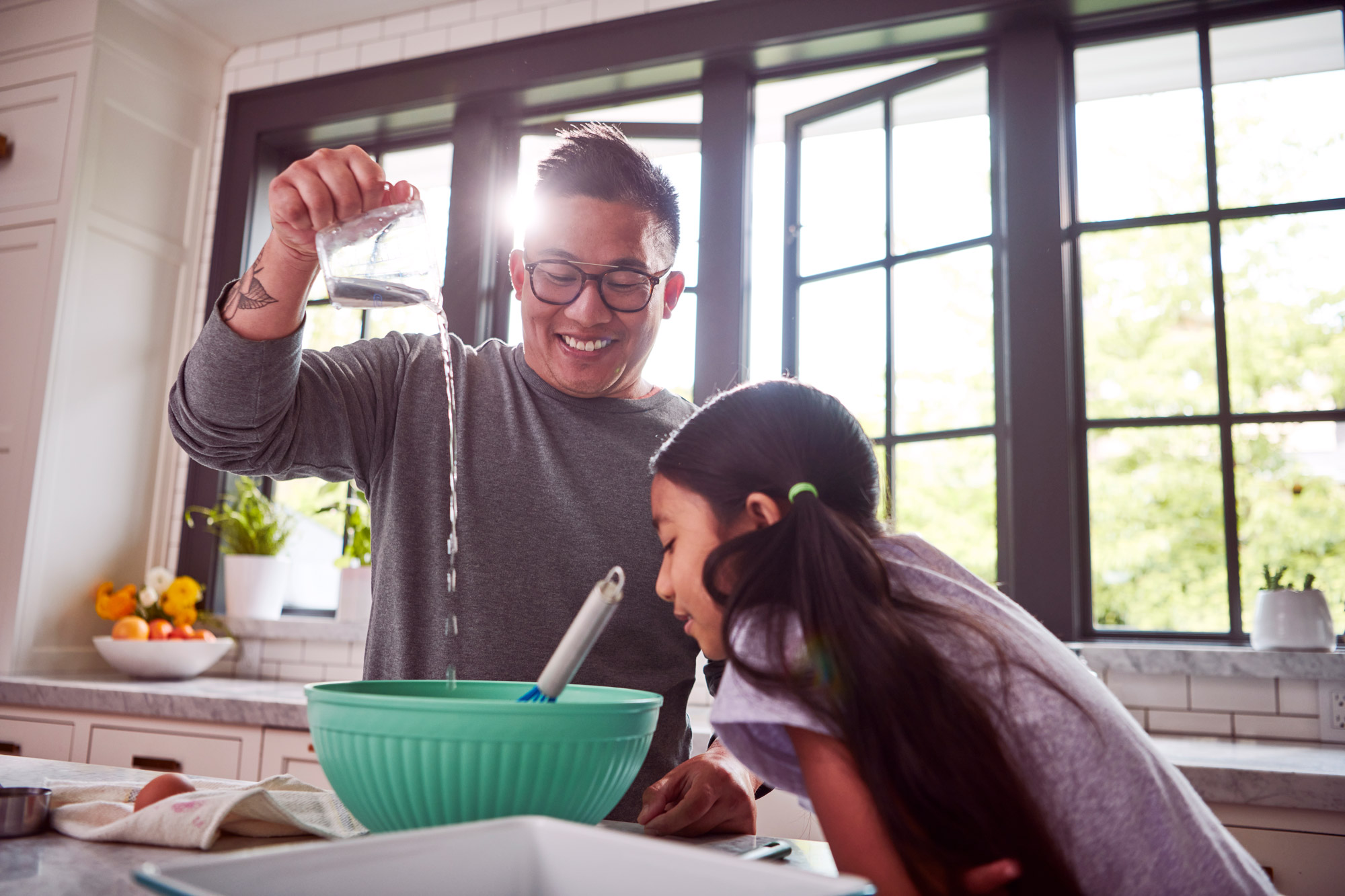 A dad pours water into a green mixing bowl while his daughter watches.